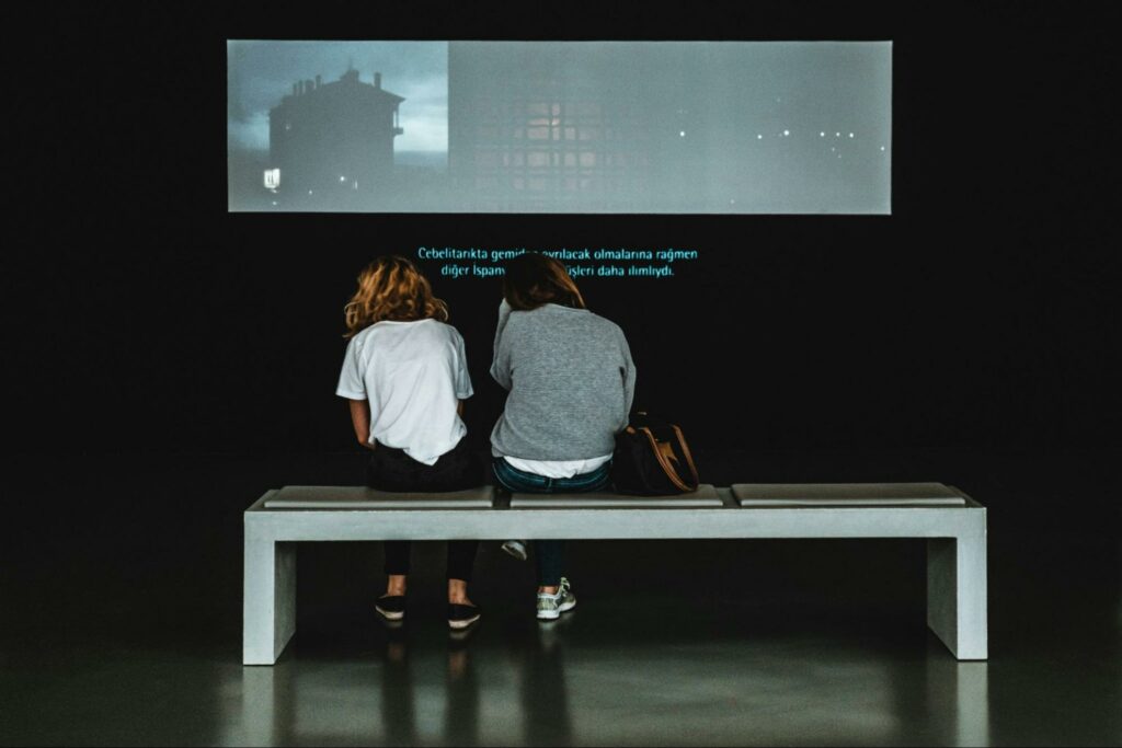 Two people sitting on a bench in a gallery watching a video with translated subtitles